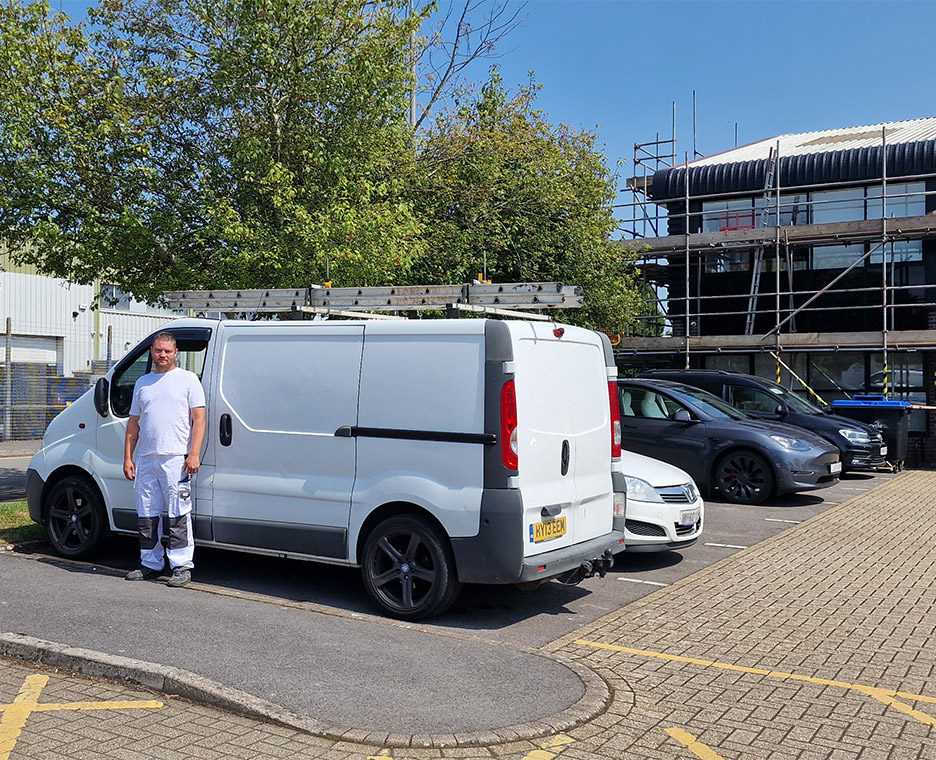 Worker standing next to his van. A shiny large commercial building with a new paint coat is shown in the background.